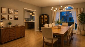 Un coin repas avec une table et des chaises en bois, une cuisine à l’arrière-plan et une grande fenêtre avec des rideaux.