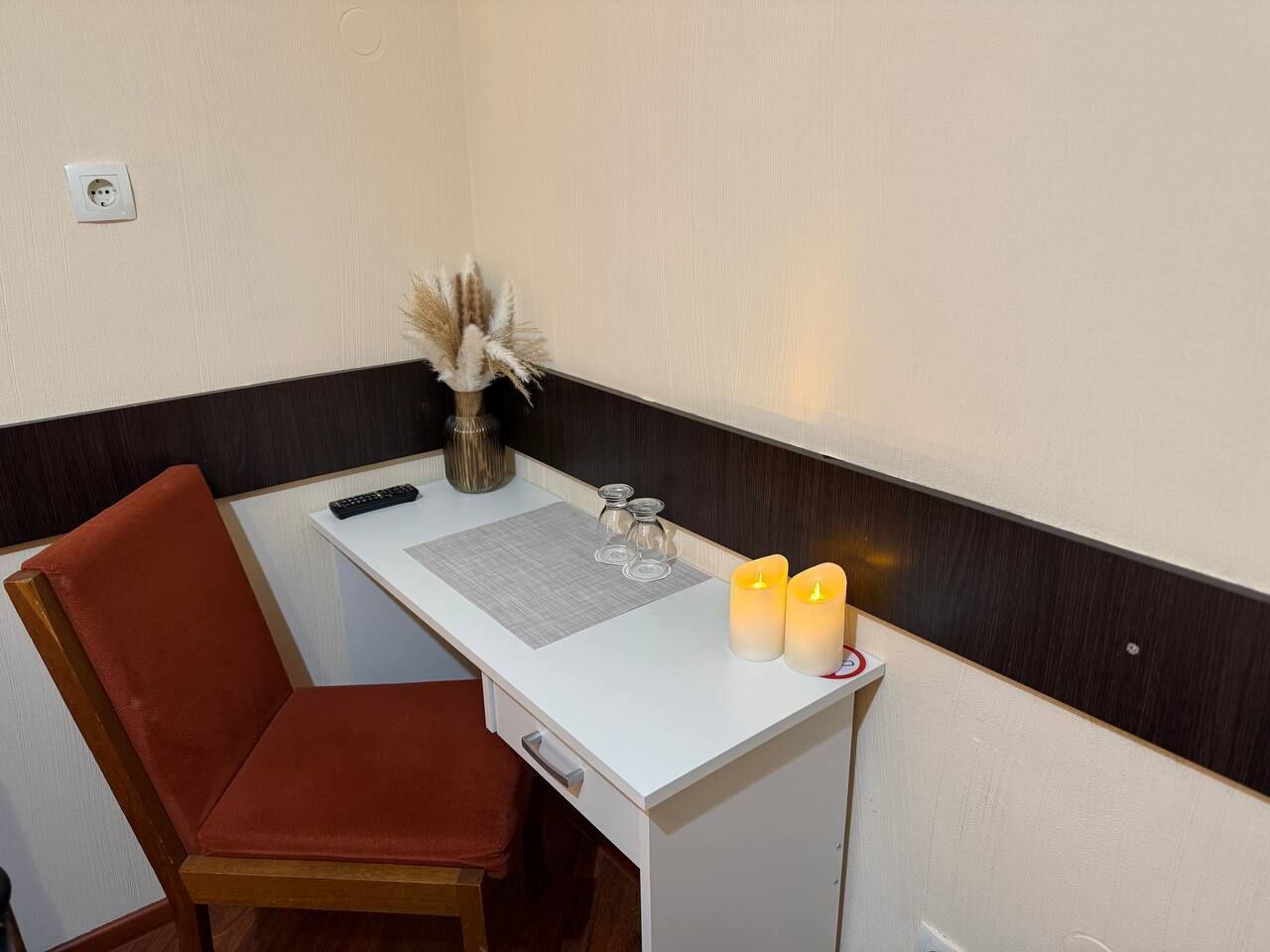 Een klein, afgescheiden kantoor met een bureau, stoel en een vaas met gedroogde planten.