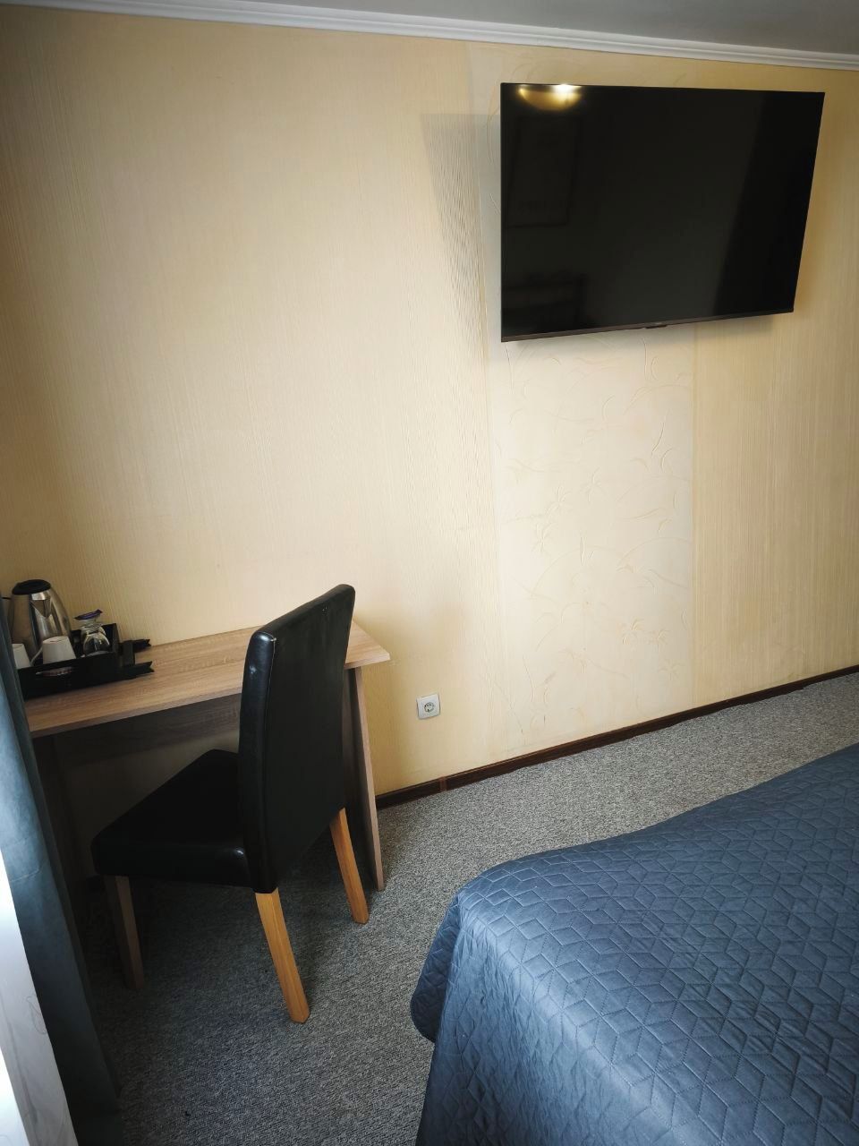 A hotel room with a bed, a desk with a chair, a flat-screen TV mounted on the wall, and a small table with a coffee maker.