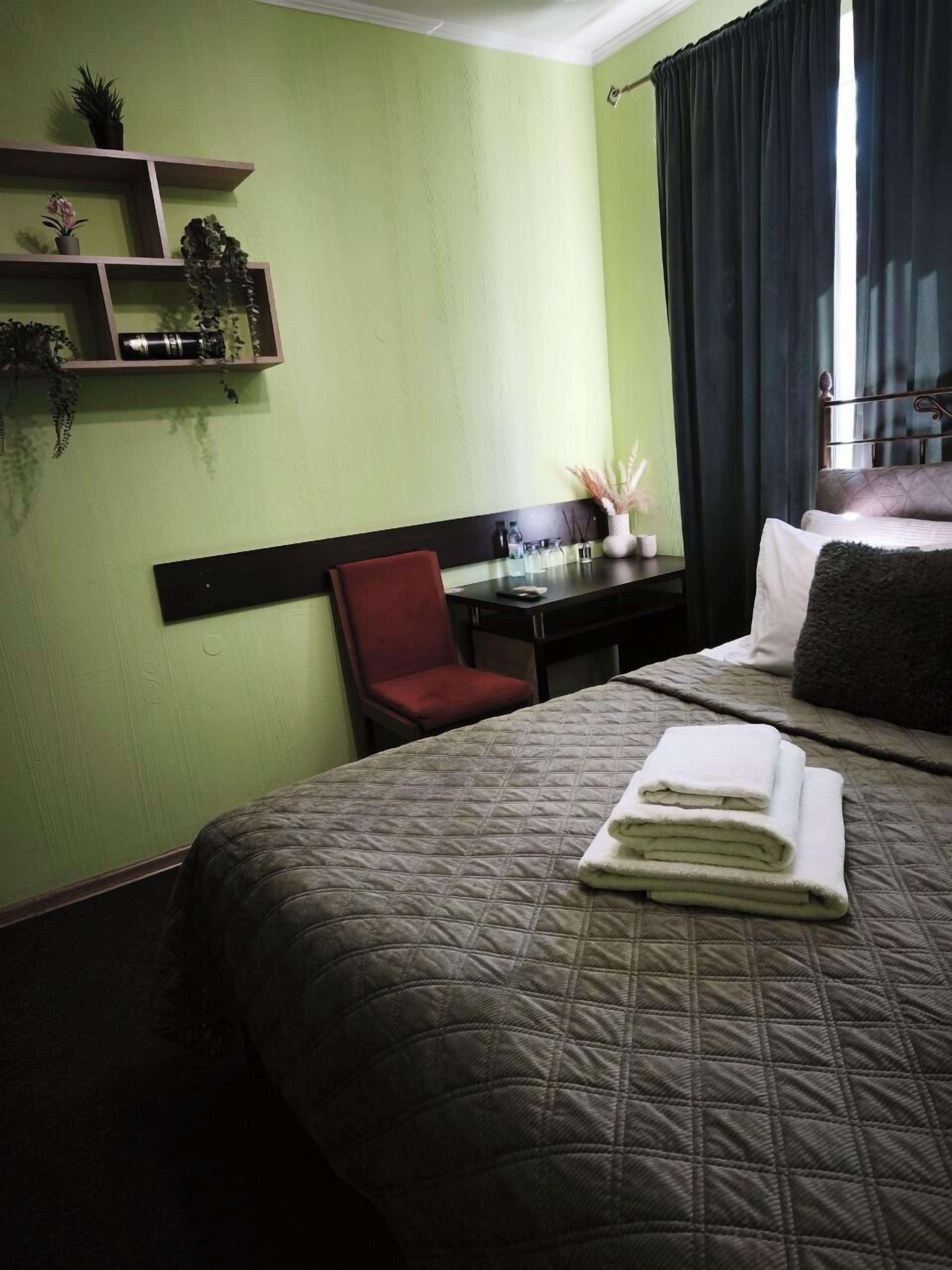 A hotel room with a bed, a desk, a chair, and a shelf with plants.