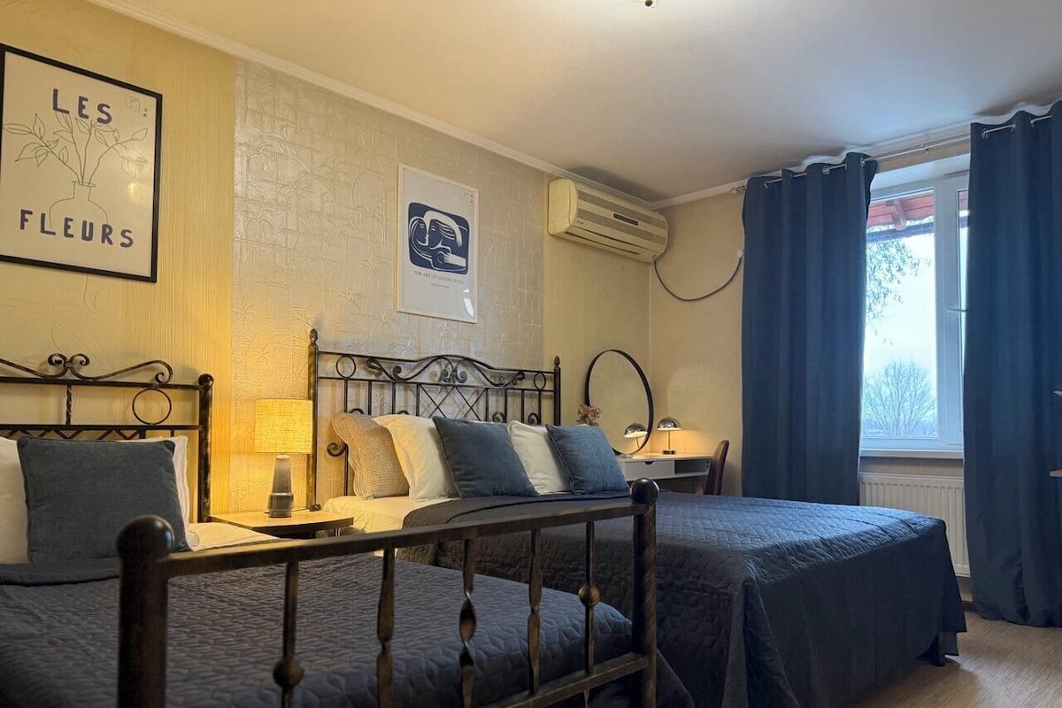 A bedroom with two beds, a bedside table with a lamp, a wall-mounted air conditioner, and a window with curtains.