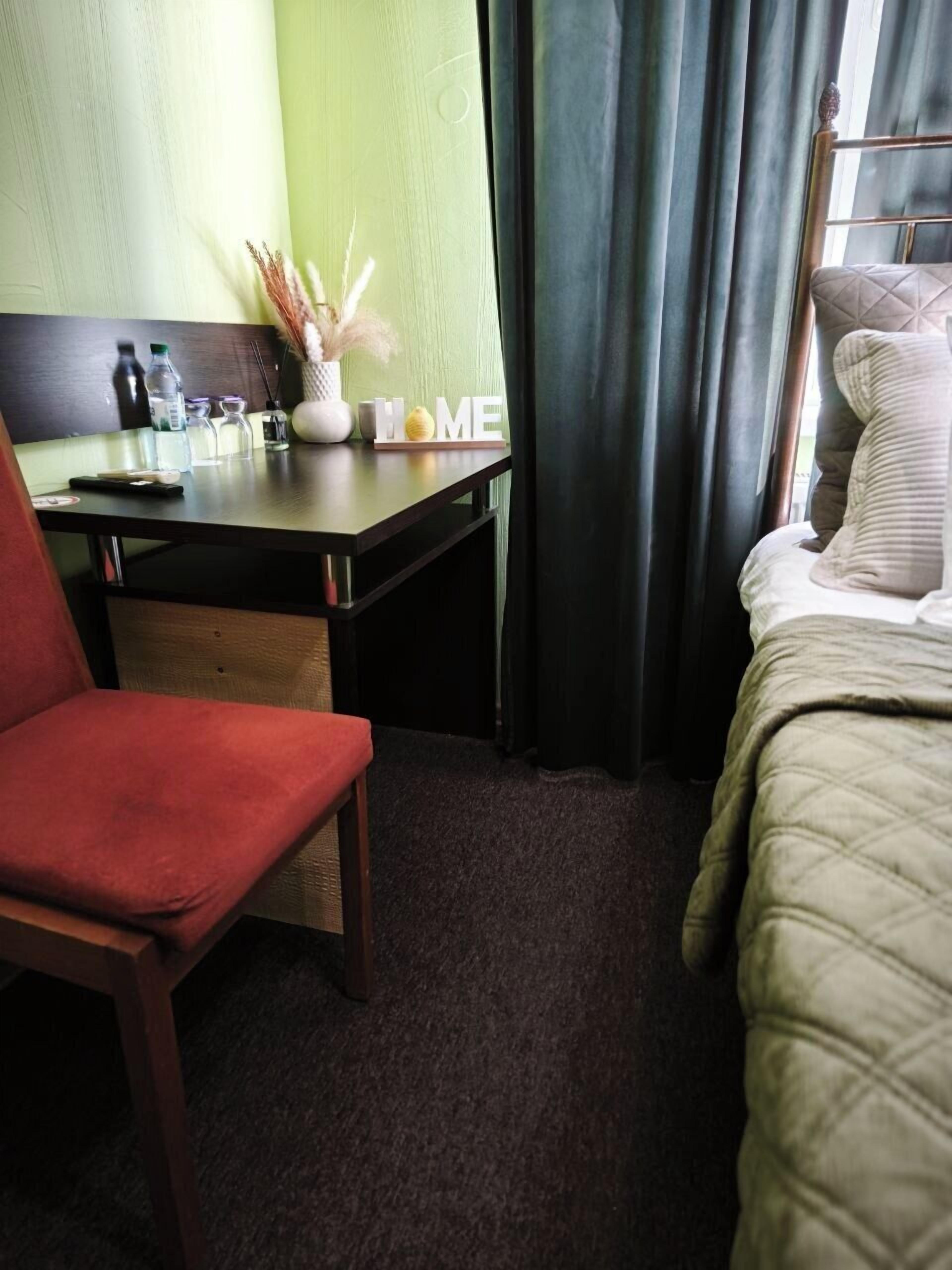 Habitación de hotel con cama, escritorio y silla, una mesita con un florero y un letrero que dice 'HOME'.