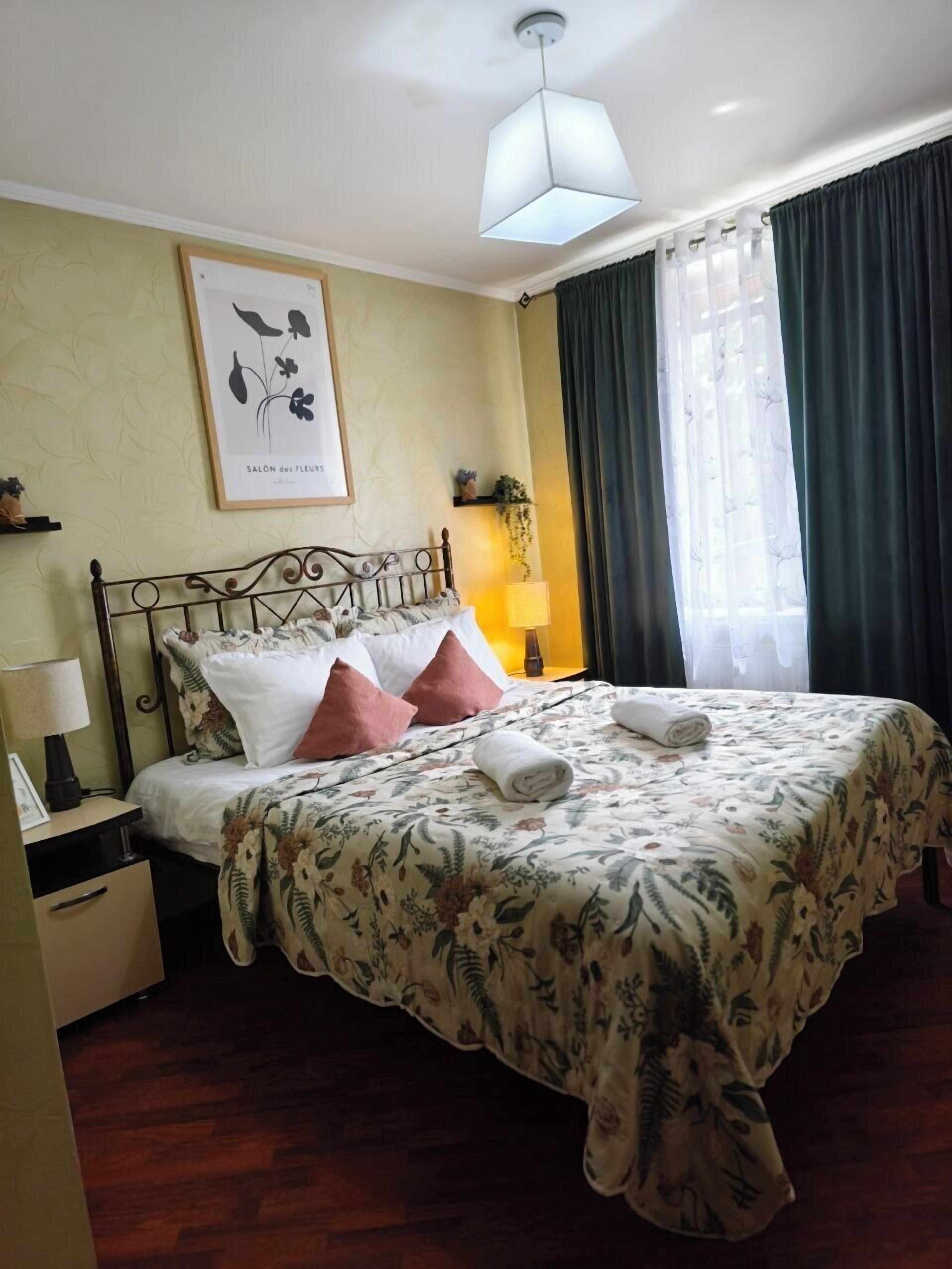 A neatly made bed with floral bedding, a bedside lamp, a framed floral artwork on the wall, and a window with curtains.