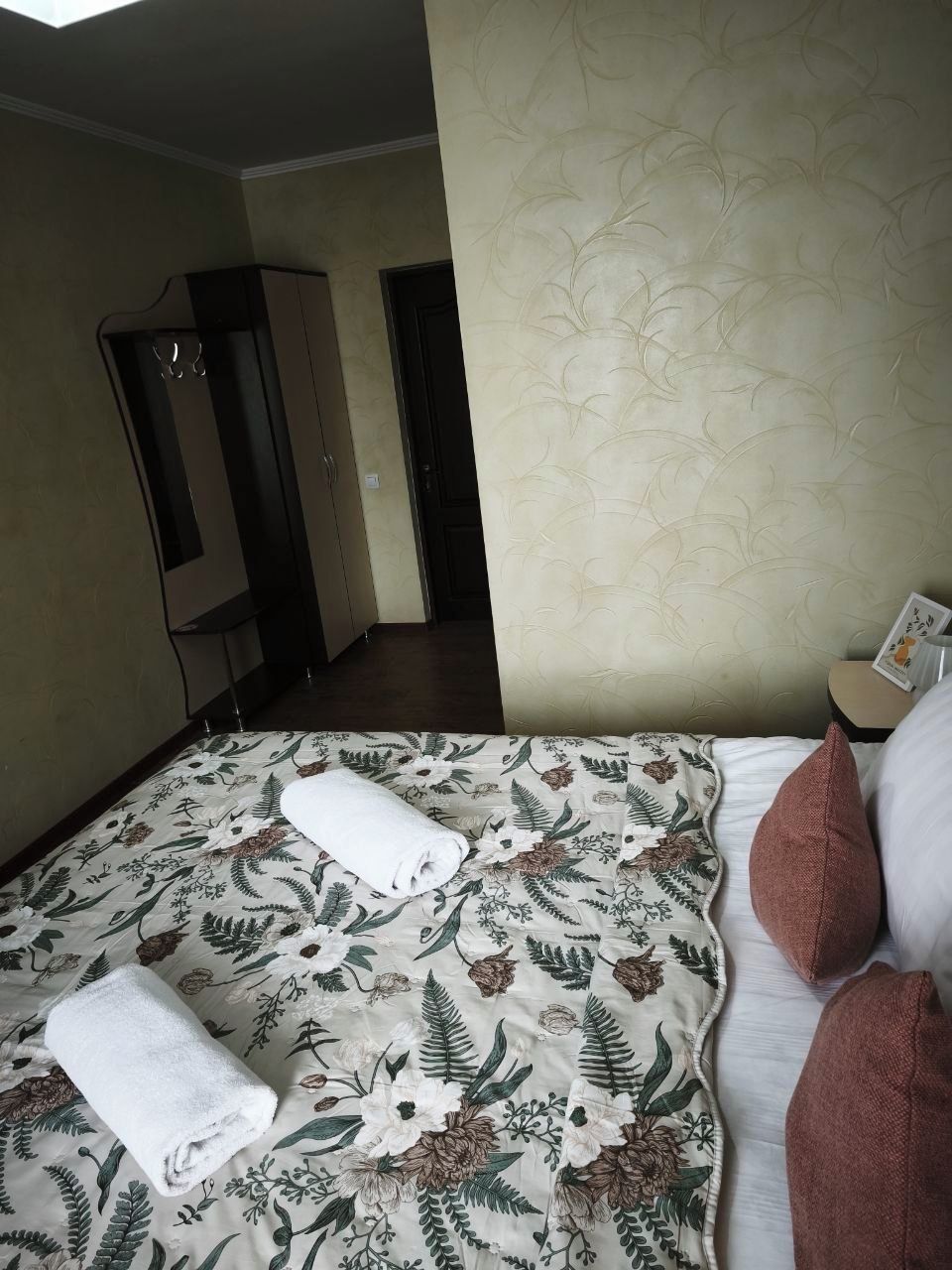 A hotel room with a bed, two rolled towels, a nightstand, and a wardrobe.