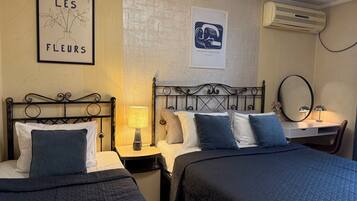 A hotel room with two beds, a desk, and a chair. There are wall-mounted air conditioning units and framed artwork on the walls.