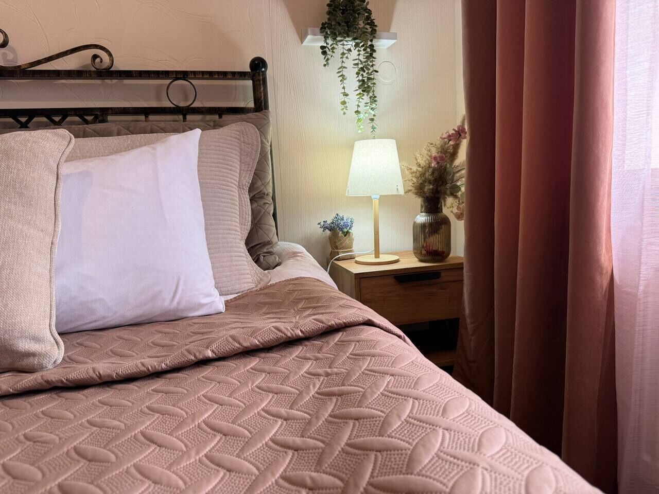 Ein ordentlich bezogenes Bett mit einer gesteppten Decke, ein Nachttisch mit einer Lampe und eine hängende Pflanze.