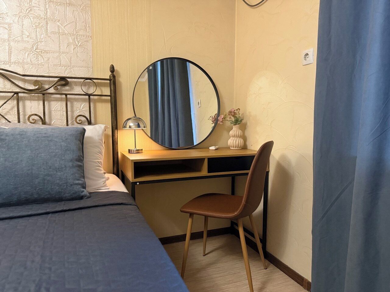 A bedroom with a bed, a desk, a chair, a round mirror, and a lamp.