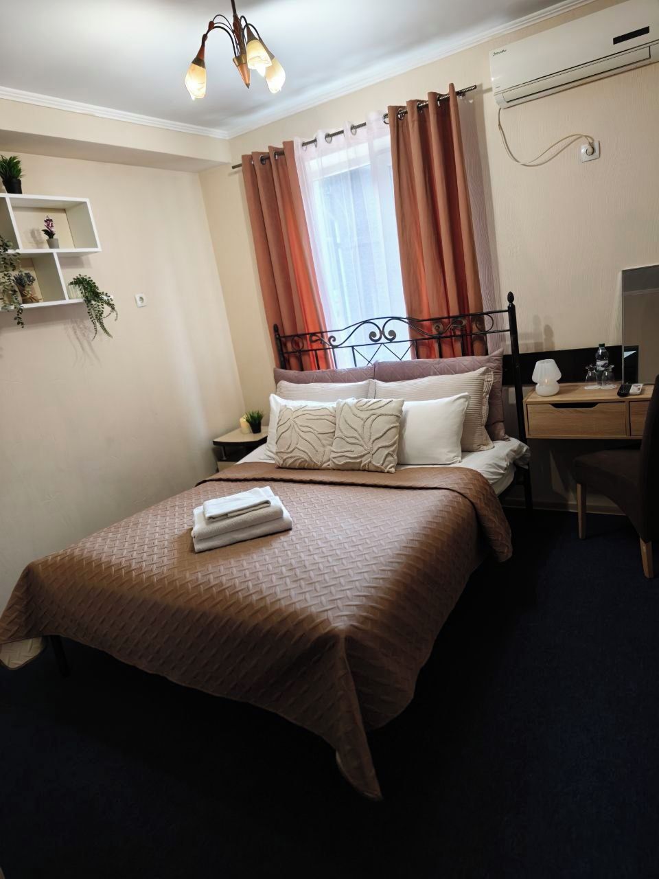 A hotel room with a bed, a desk, a chair, a nightstand, a wall-mounted air conditioner, and a ceiling light fixture.