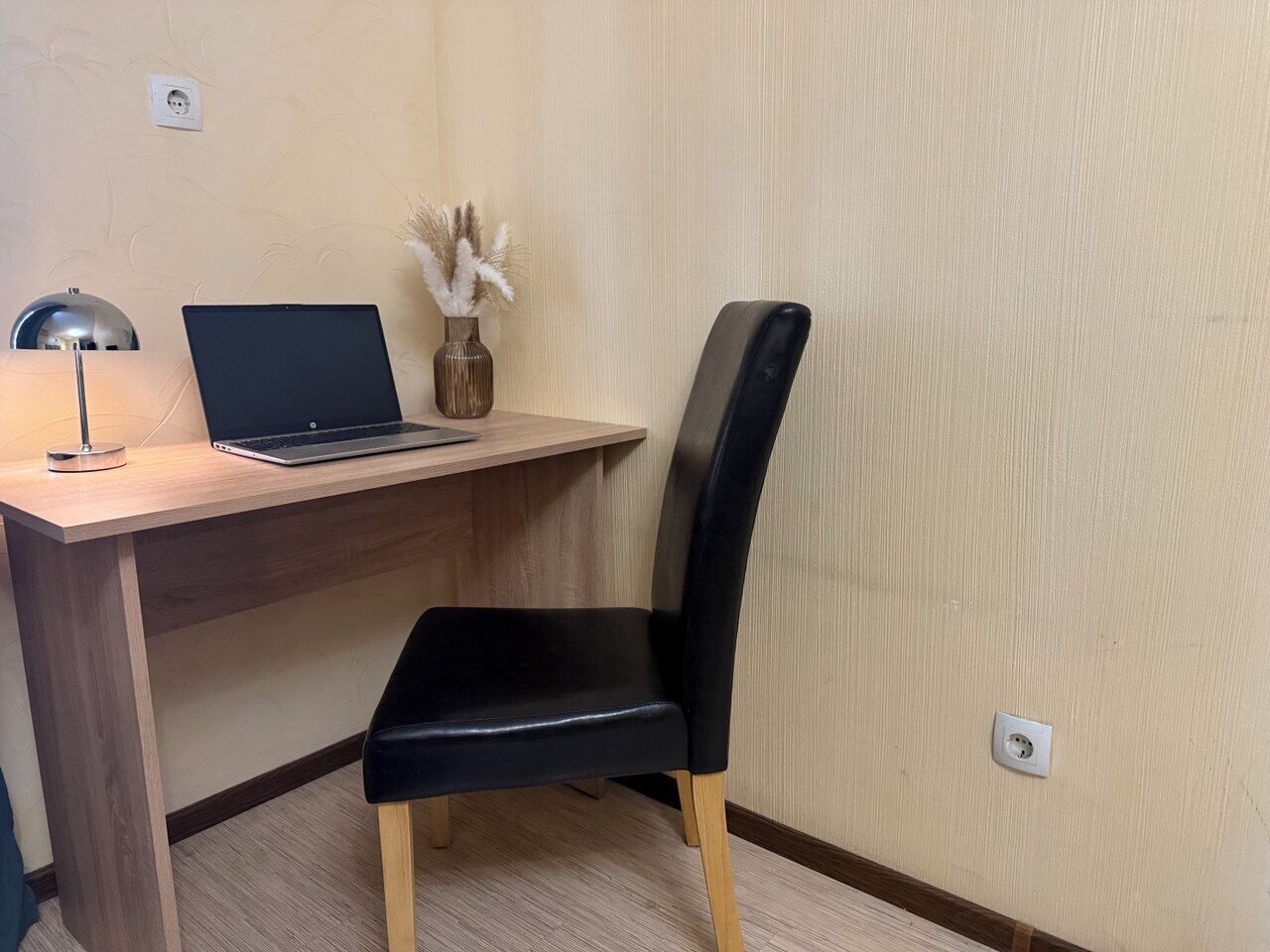 A workspace with a wooden desk, a black chair, a laptop, a vase with dried plants, and a desk lamp.