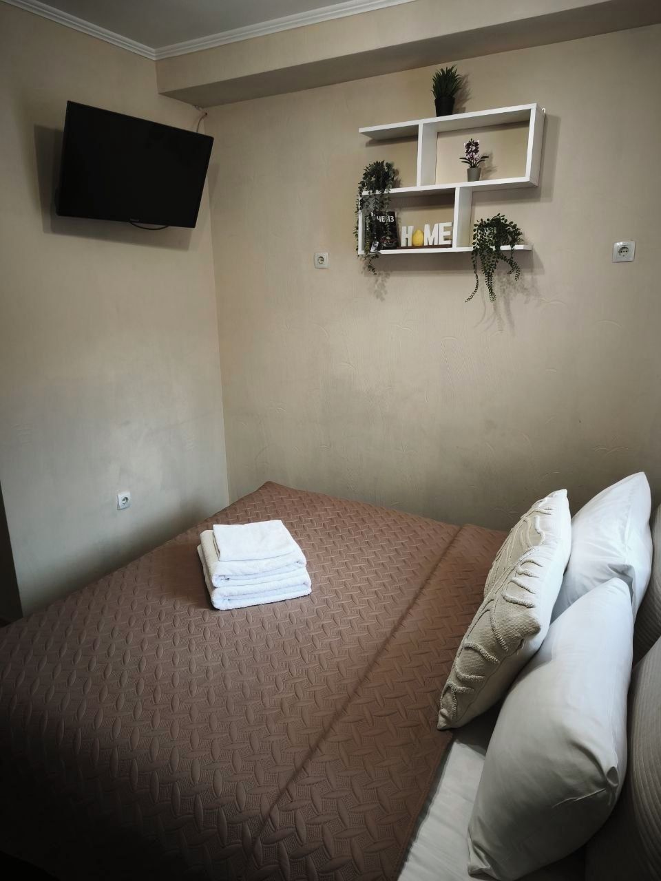 Une chambre à coucher avec un lit, des oreillers, une télévision et une étagère ornée de plantes et d’une décoration portant l’inscription « HOME ».