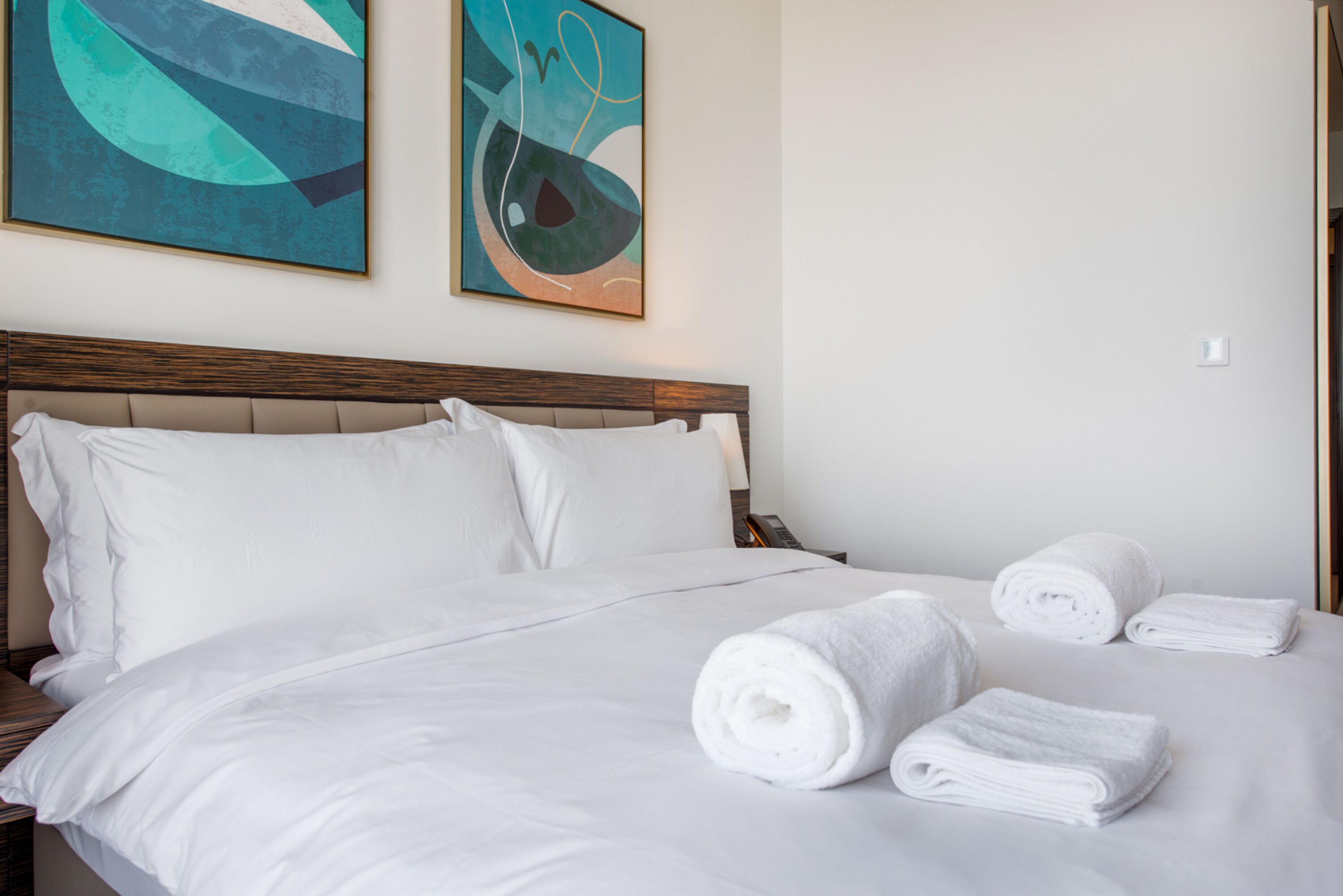 Een net opgemaakt bed met witte lakens en opgerolde handdoeken, een houten hoofdbord en abstracte kunst aan de muur.