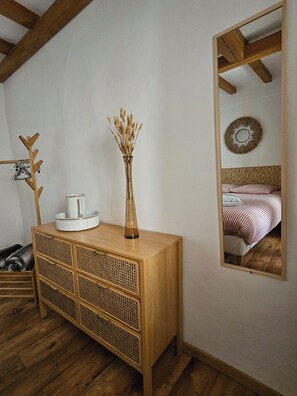 Une commode en bois avec un miroir, un vase contenant des plantes séchées et un miroir mural qui reflète un lit avec une literie à rayures.