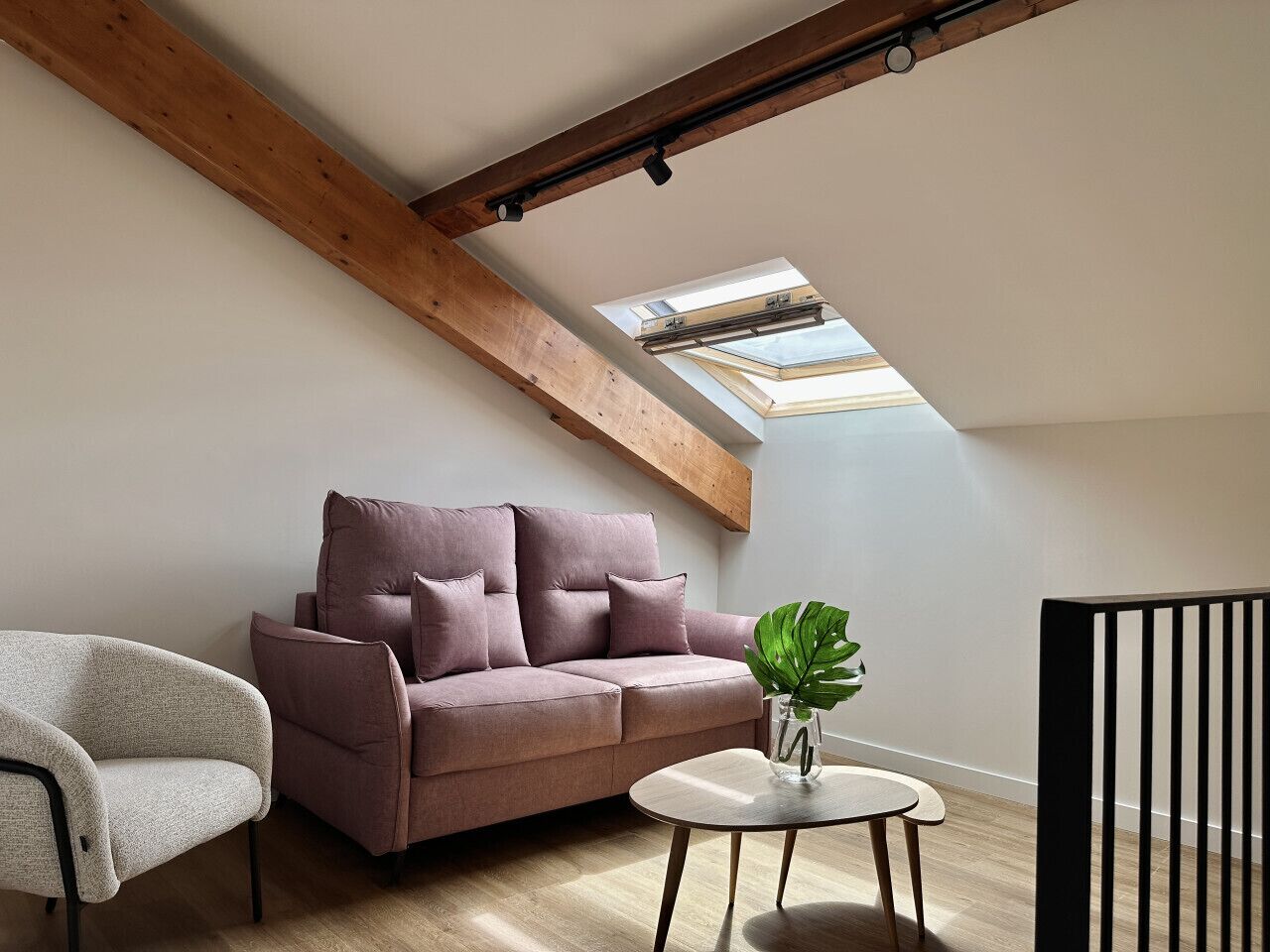 A modern loft bedroom with a sloped ceiling, a large window, a sofa, a chair, and a small table with a plant.