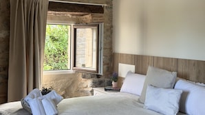 A bedroom with a stone wall, a large window with curtains, a bed with white bedding, and a bedside table with books and a vase of flowers. - Casa De Postas - Duplex Superior (San Miguel de Meruelo)