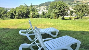 Twee witte plastic ligstoelen op een gazon, met uitzicht op een dorp en heuvels op de achtergrond.