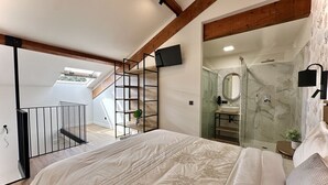 Een slaapkamer met een groot bed, een aan de muur bevestigde televisie, een badkamer met een douchecabine en een trap naar een hoger gelegen verdieping.