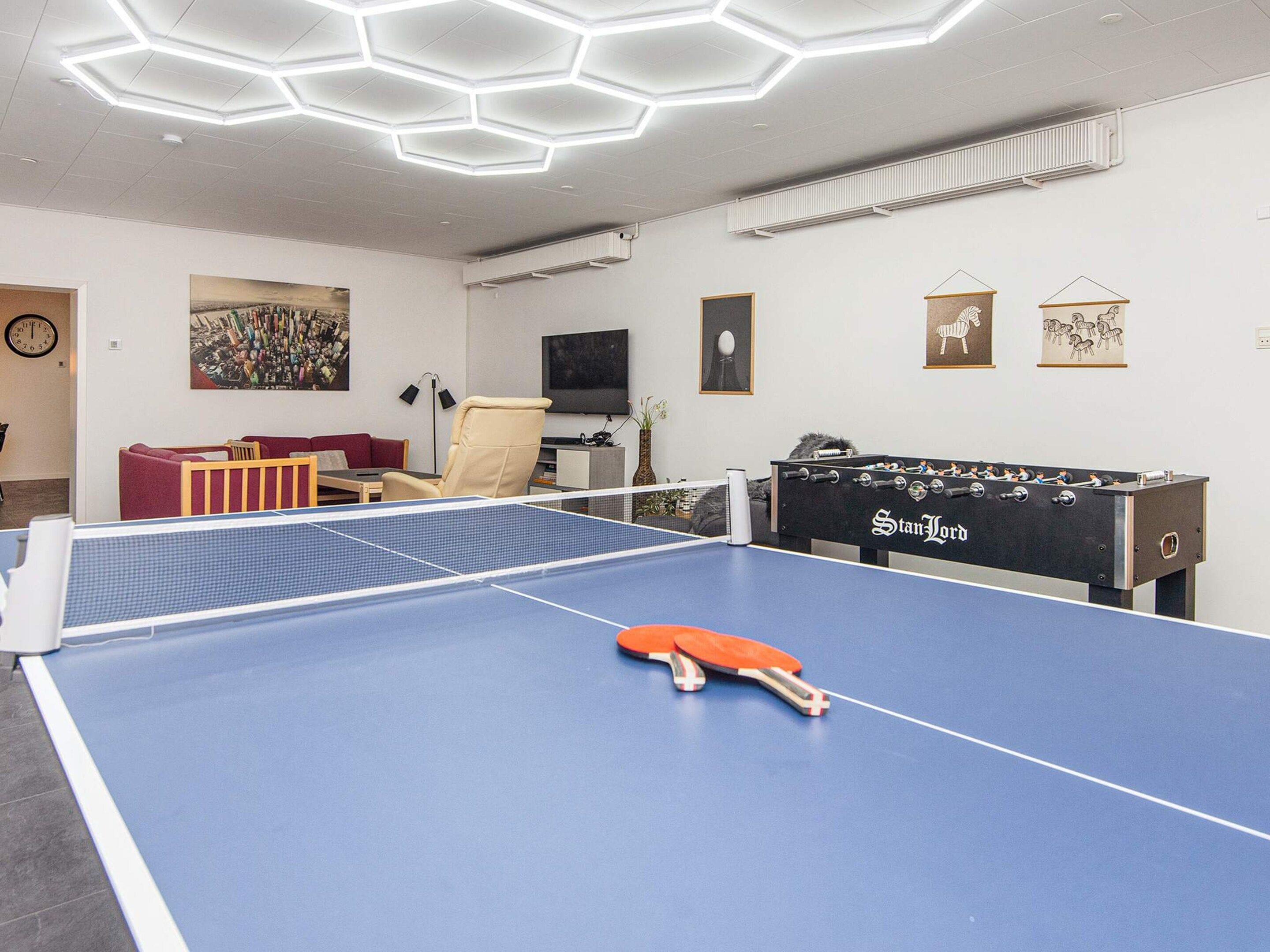A room with a ping pong table, a foosball table, a sofa, a chair, and artwork on the walls.