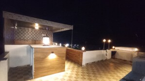 Een terras op het dak met een geruit vloertje, een bar met houten details en meerdere lampen.