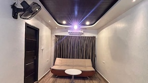 Een kleine kamer met een bank en een tafeltje, een plafondventilator en een airconditioning.