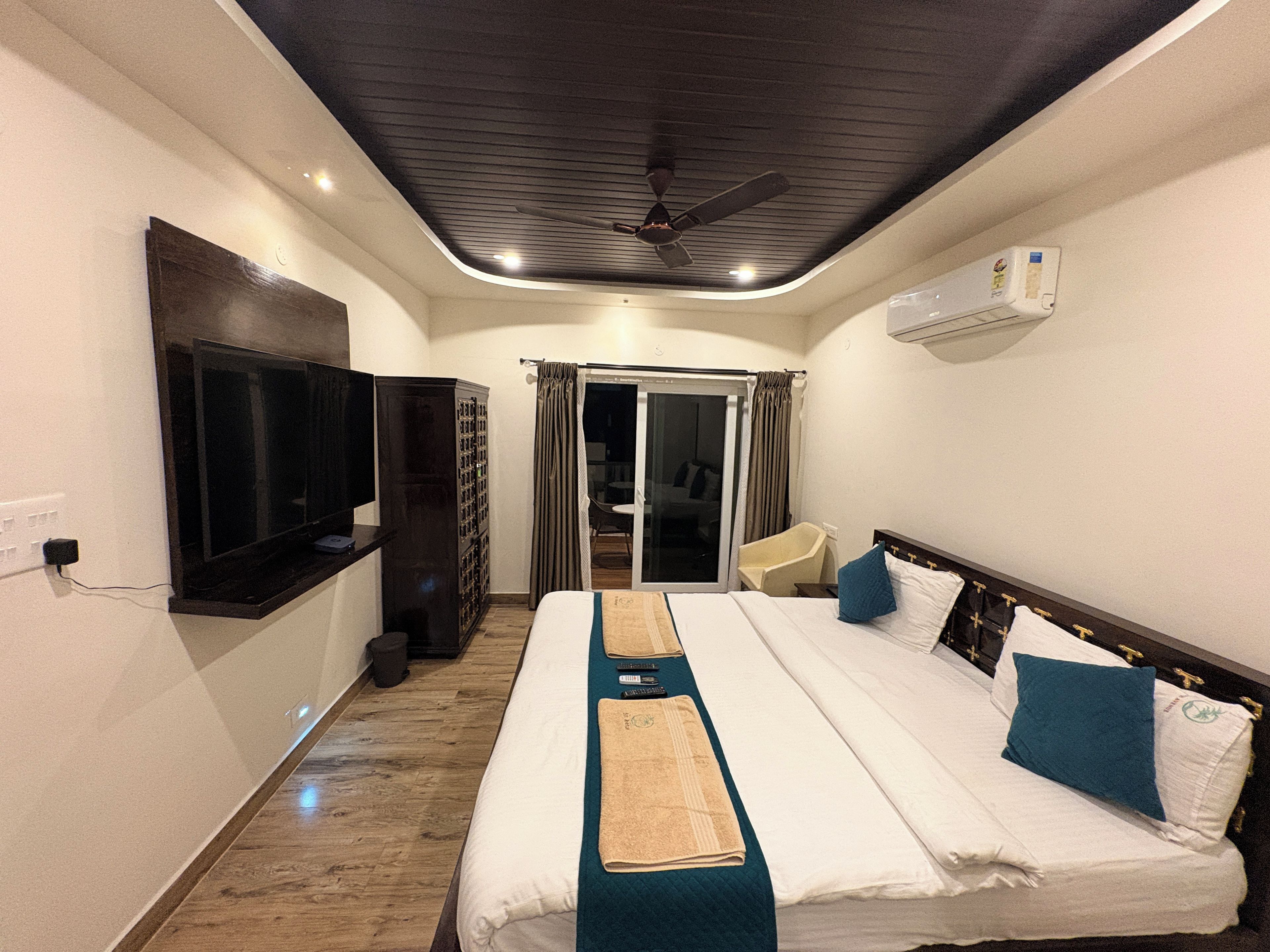 Una habitación de hotel con cama, televisión, ventilador de techo y puerta corredera que da a un balcón.