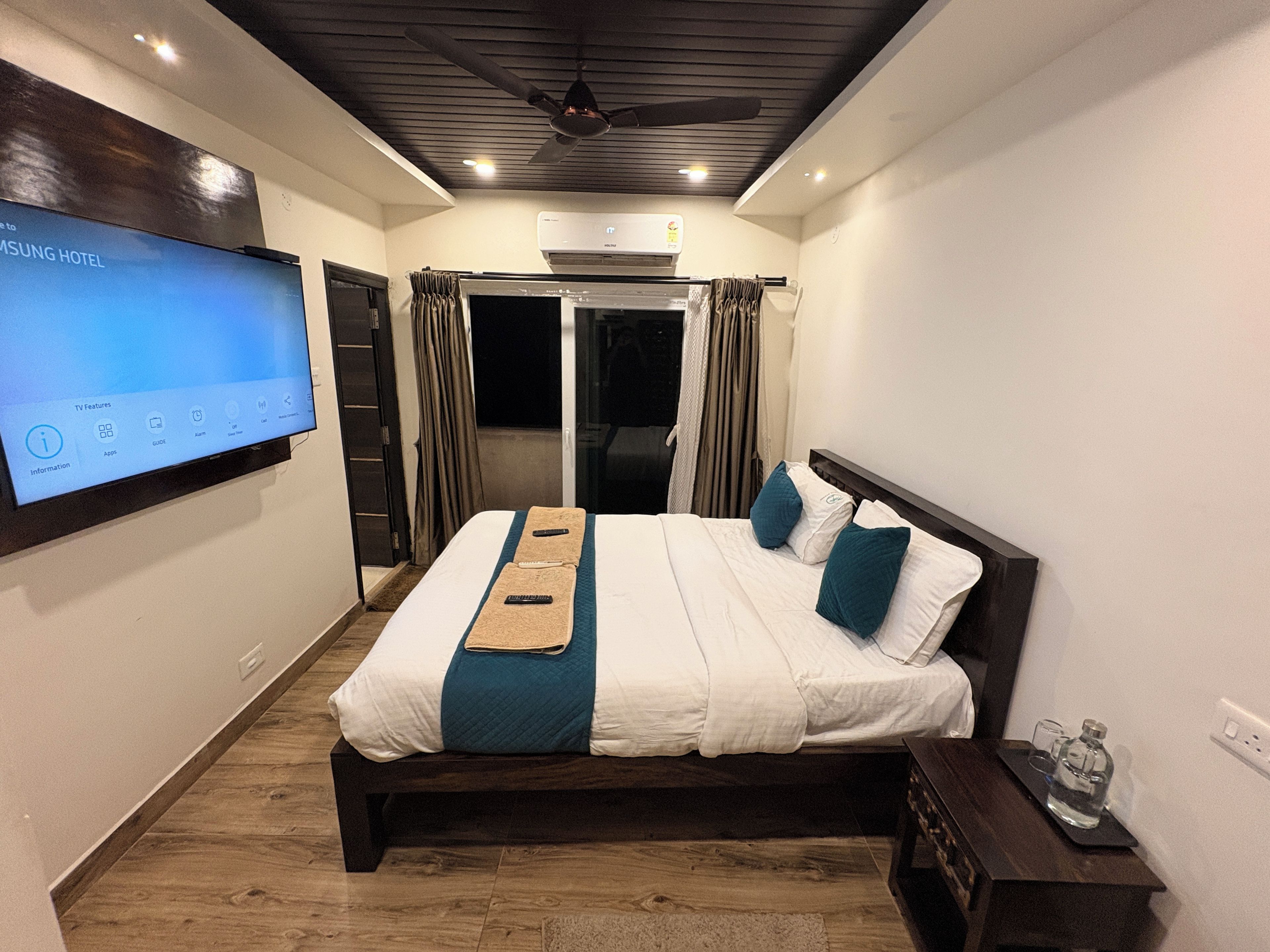 Ein Hotelzimmer mit einem Bett, einem an der Wand montierten Fernseher, einem Deckenventilator und einer Klimaanlage.