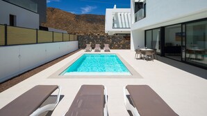 Ett modernt utomhusområde med pool, solstolar, ett matbord och en bakgrundsvägg av sten.