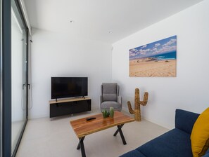 A modern living room with a flat-screen TV, a grey armchair, a wooden coffee table, a cactus sculpture, and a blue sofa with a yellow pillow. - Deluxe Las Calderas 01 With Private Pool (La Oliva)