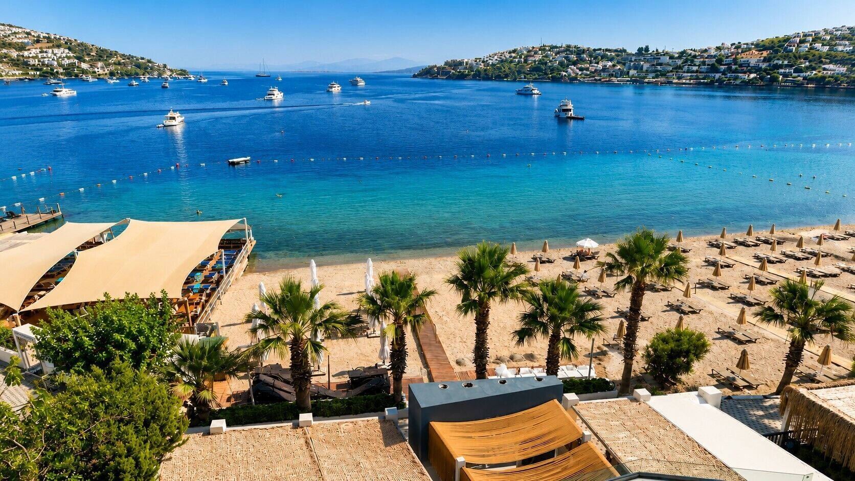 Una playa con una hilera de palmeras, arena dorada y vistas a barcos en el agua.