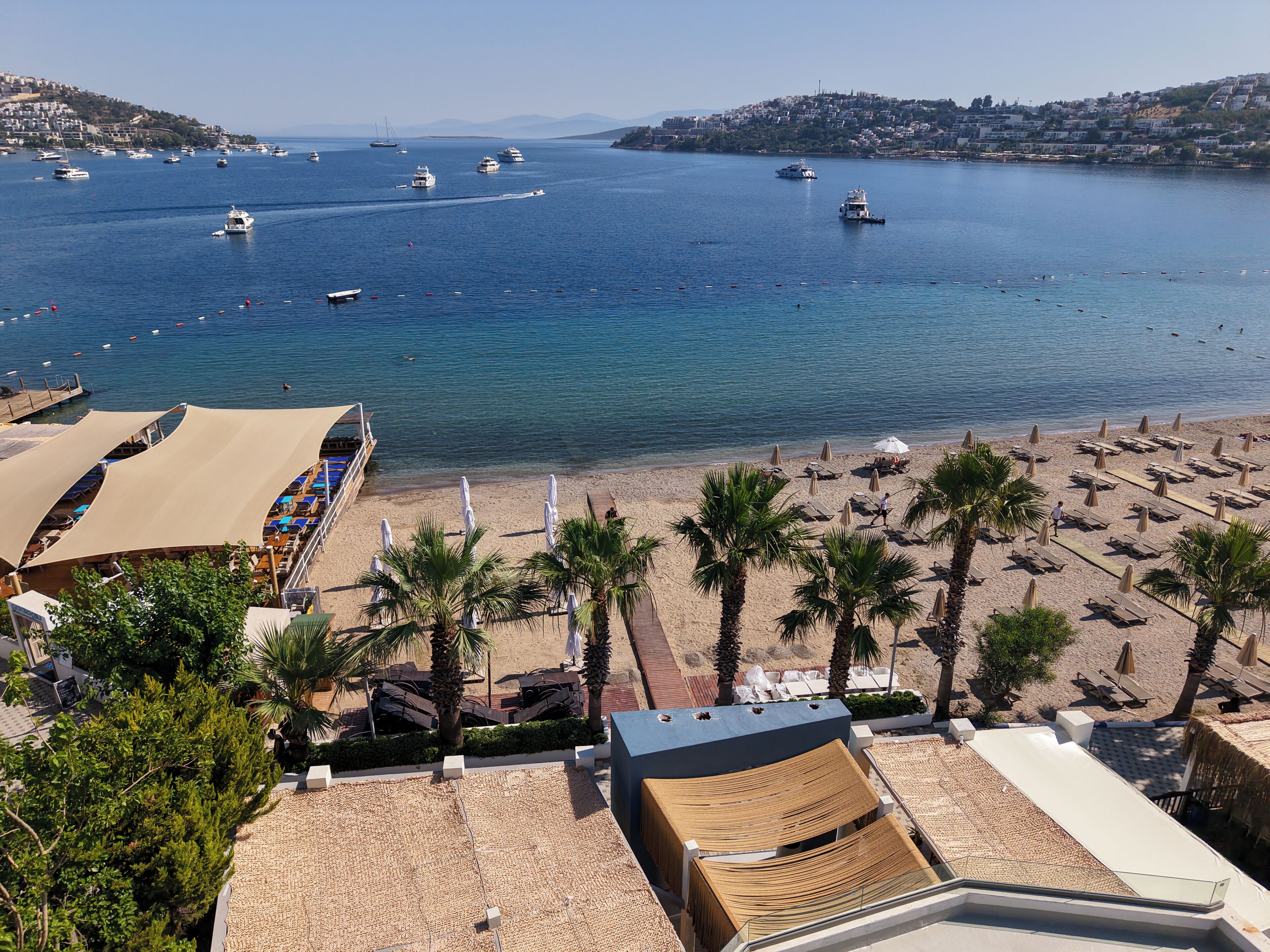 A beach with umbrellas and lounge chairs, a view of the sea with boats, and a hillside in the background.