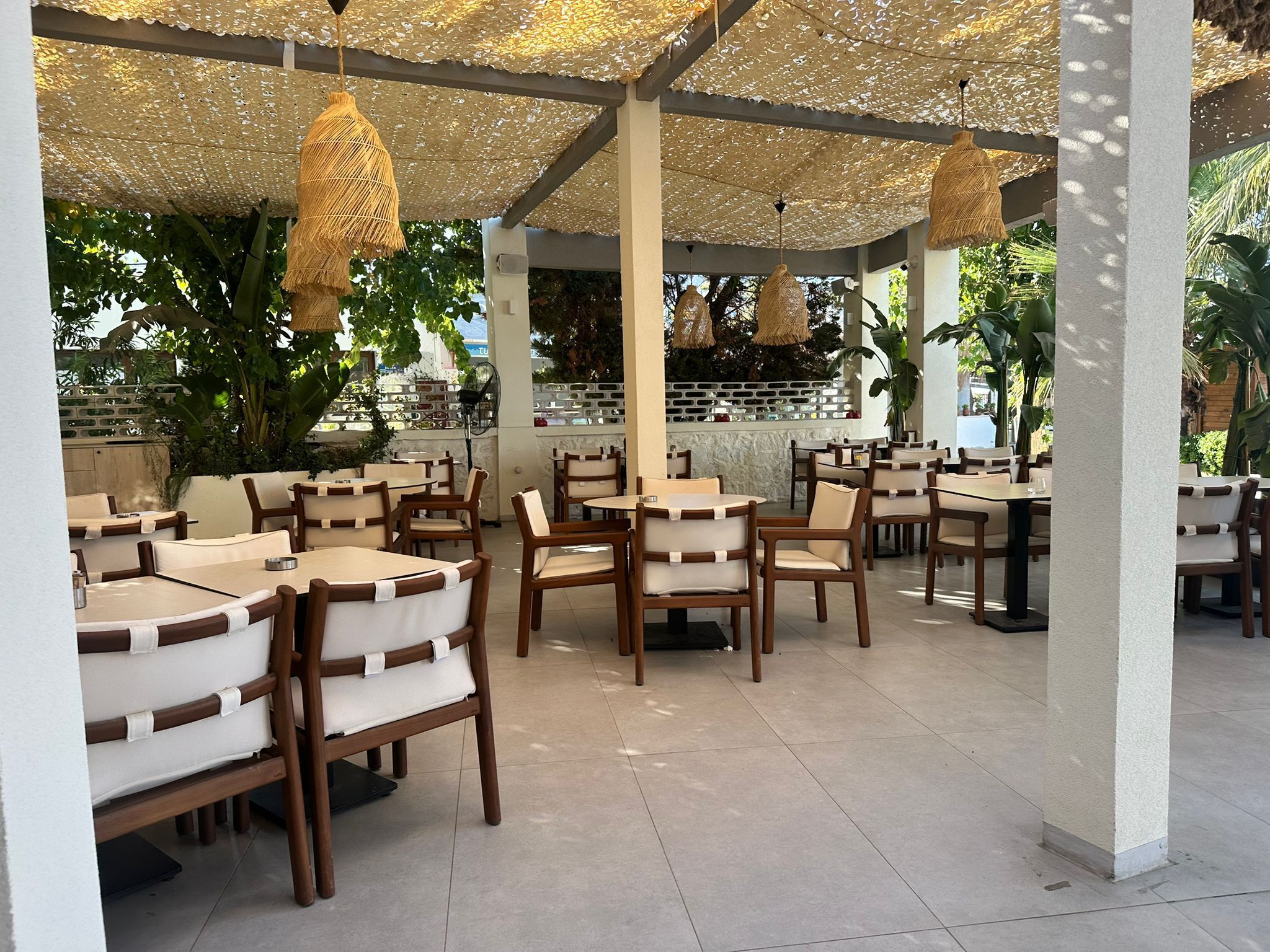 Un restaurante al aire libre con mesas y sillas de madera, un muro de ladrillo y lámparas colgantes de mimbre.