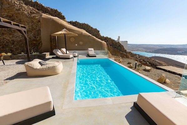 Una piscina con sillones de descanso y una pérgola, rodeada de un paisaje rocoso y vista al mar.