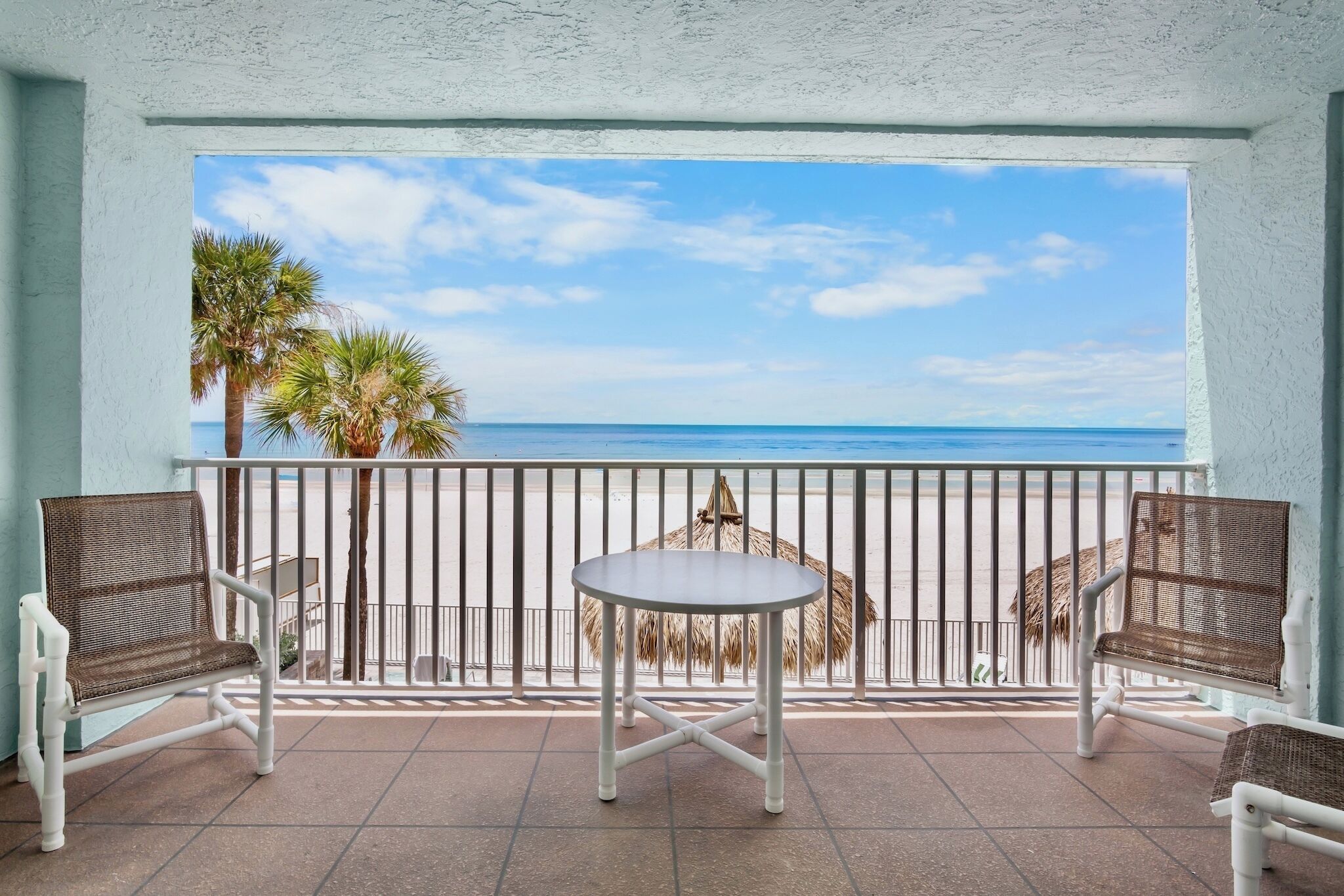 A balcony with two wicker chairs and a small round table, overlooking a beach with palm trees and a clear sky.