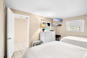 Un dormitorio con dos camas, un aparador, un banco, un escritorio, una lámpara y un televisor montado en la pared.