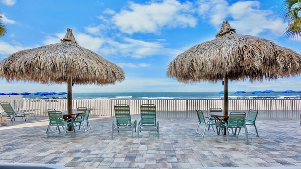 A beachside patio with thatched umbrellas, green chairs, and a view of the ocean.