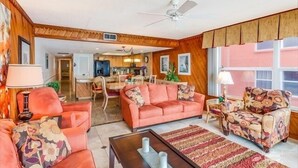 A living room with a sofa, armchair, and coffee table. There is a ceiling fan and a window with curtains.