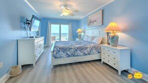 A bedroom with a bed, two nightstands, a ceiling fan, a TV mounted on the wall, and a window with curtains.