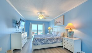 Ein Schlafzimmer mit einem Bett, zwei Nachttischen, einem Deckenventilator, einem an der Wand montierten Fernseher und einem Fenster mit Vorhängen.
