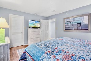 A bedroom with a bed, a television on a dresser, a window with a view of a beach, and a door.