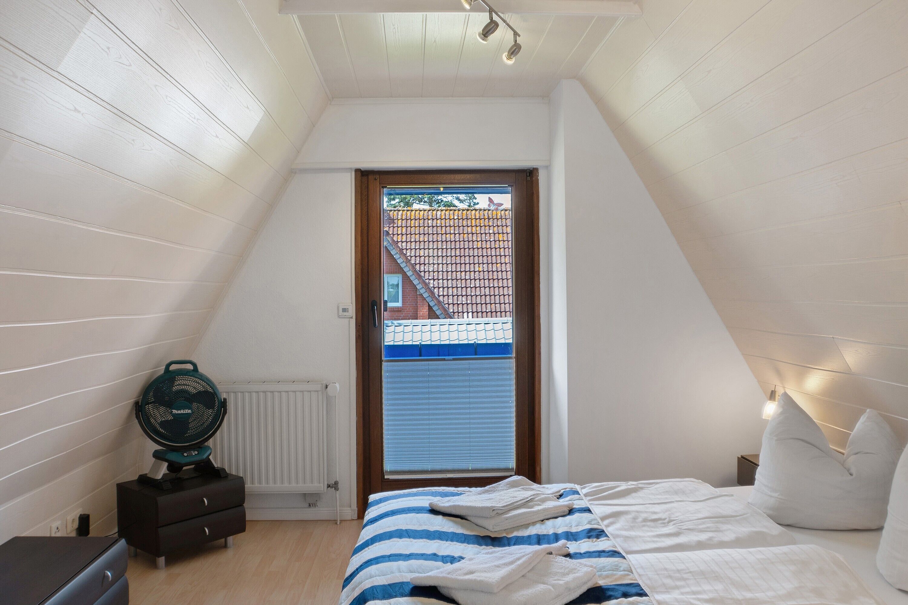 Quarto com teto inclinado, cama com roupa de cama branca e azul, mesa de cabeceira com um ventilador e janela com vista para um prédio de tijolos.