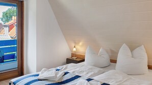 Een slaapkamer met een schuine kap, een bed met wit en blauw beddengoed, een nachtkastje met een lamp en een raam met uitzicht op gebouwen.