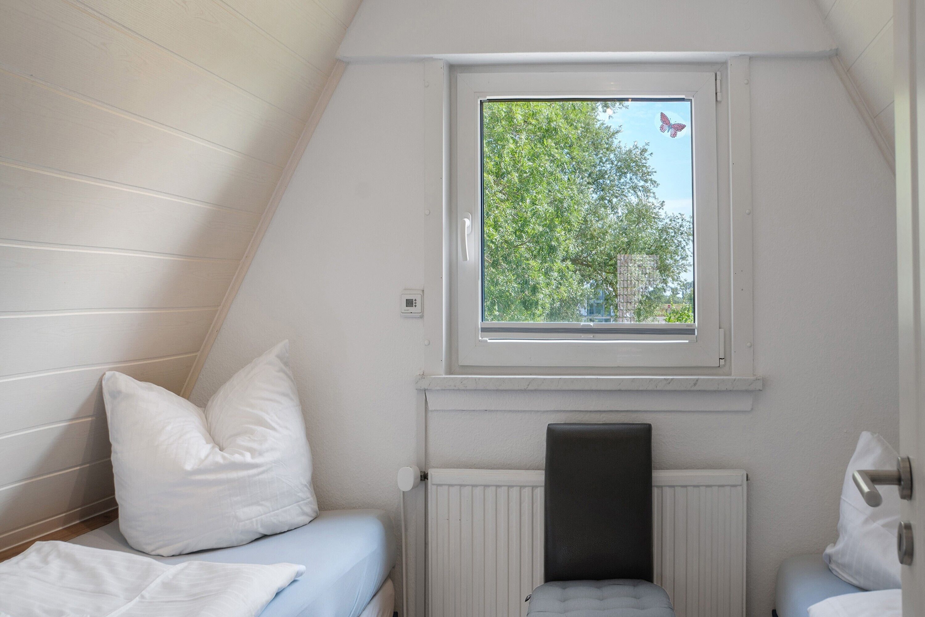 Una piccola stanza singola con un soffitto spiovente, una finestra vista alberi, un termosifone e un letto con cuscini.