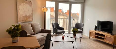 A modern living room with a sofa, two armchairs, a coffee table, a TV on a wooden stand, and a large window with a city view.