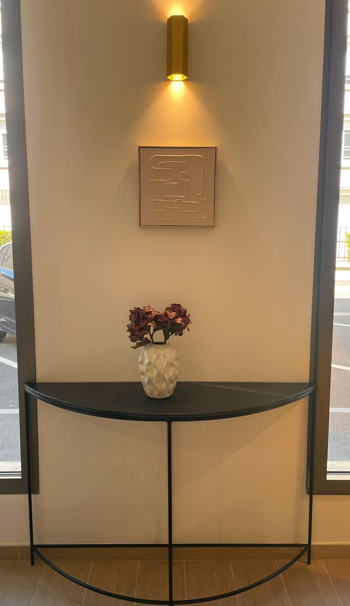 A wall-mounted light fixture, a framed abstract artwork, and a vase with flowers on a console table.