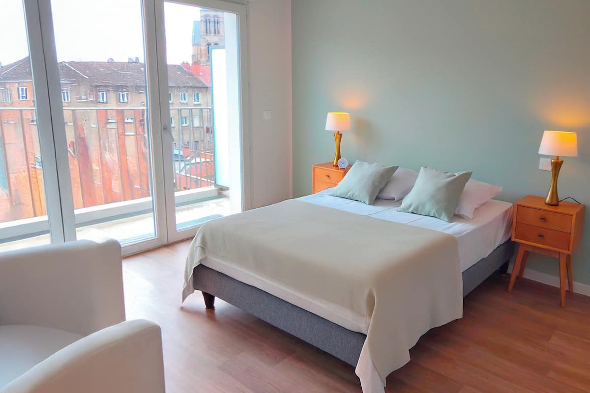 A bedroom with a bed, two bedside tables with lamps, a chair, and a large window with a view of buildings.
