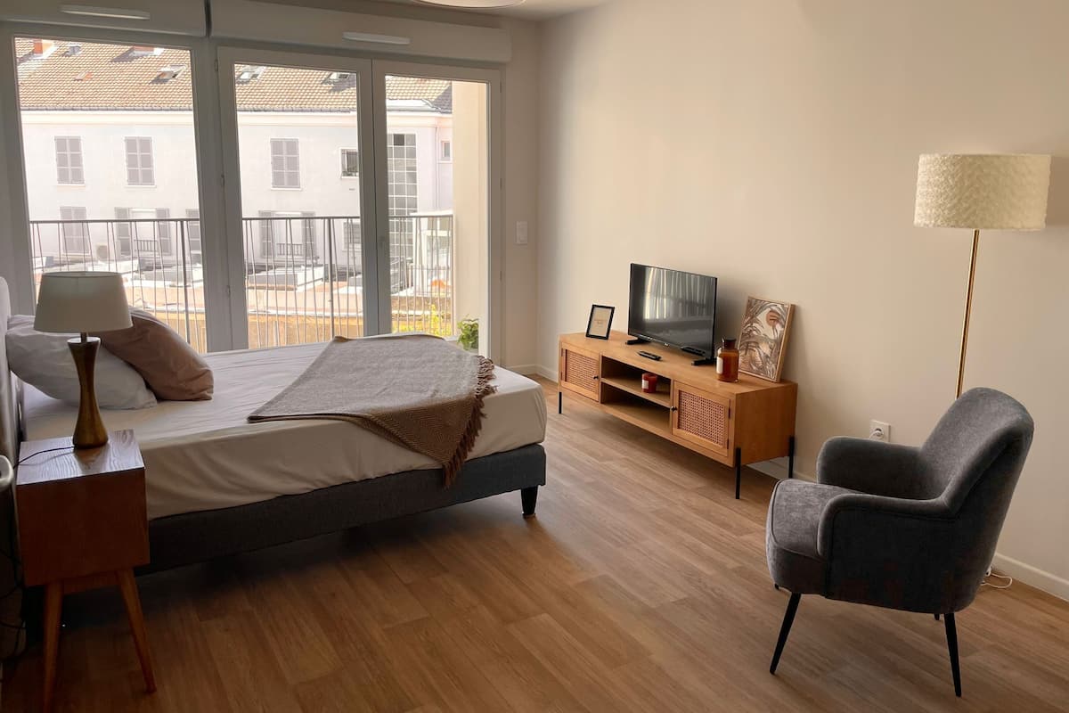 A bedroom with a bed, a TV on a wooden stand, a chair, and a balcony with a view of buildings.
