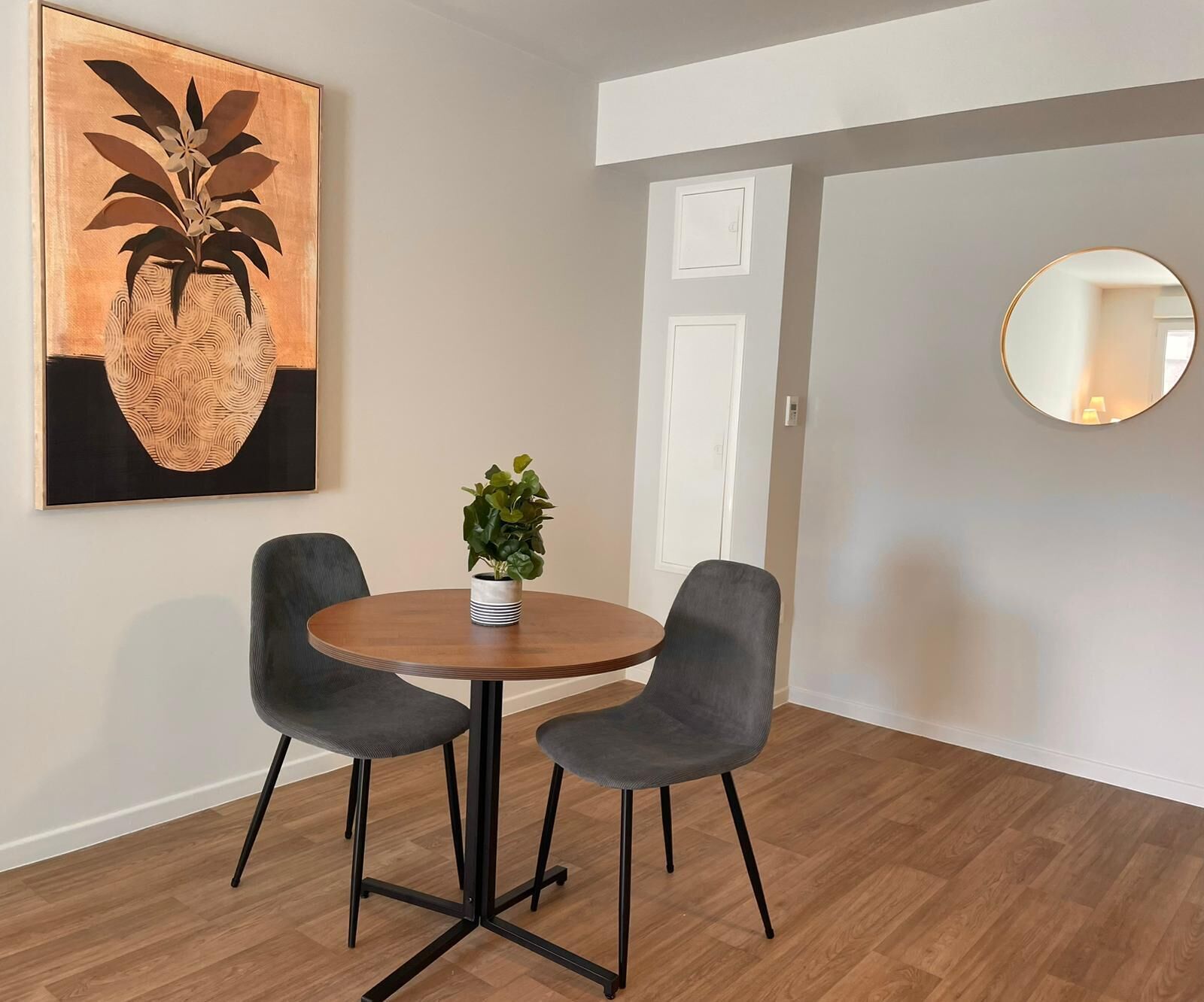 A dining area with a round table, two grey chairs, a potted plant, and a framed artwork on the wall.