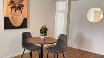 A dining area with a round table, two grey chairs, a potted plant, and a framed artwork on the wall.