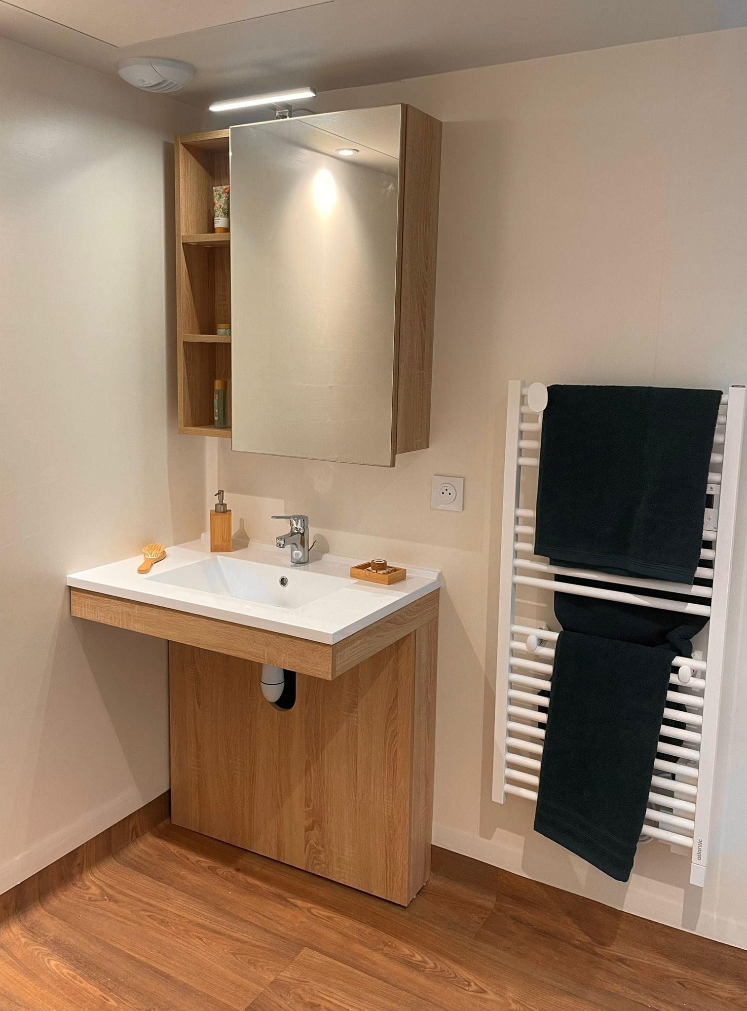 A modern bathroom with a wooden vanity, a large mirror, a white sink, and a towel rack with a dark towel.