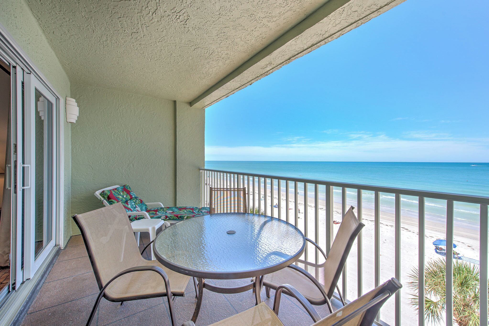 A balcony with a glass table and chairs, a view of the beach, and a door with a sliding glass window.