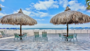 Un patio frente a la playa con sombrillas de paja, sillas verdes y vistas al océano.